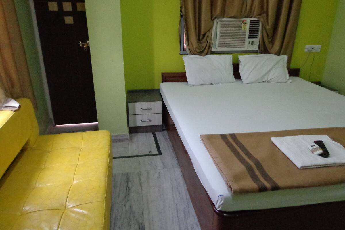 A hotel room with a bed, a bedside table, a small drawer, a yellow sofa, and a window with curtains.