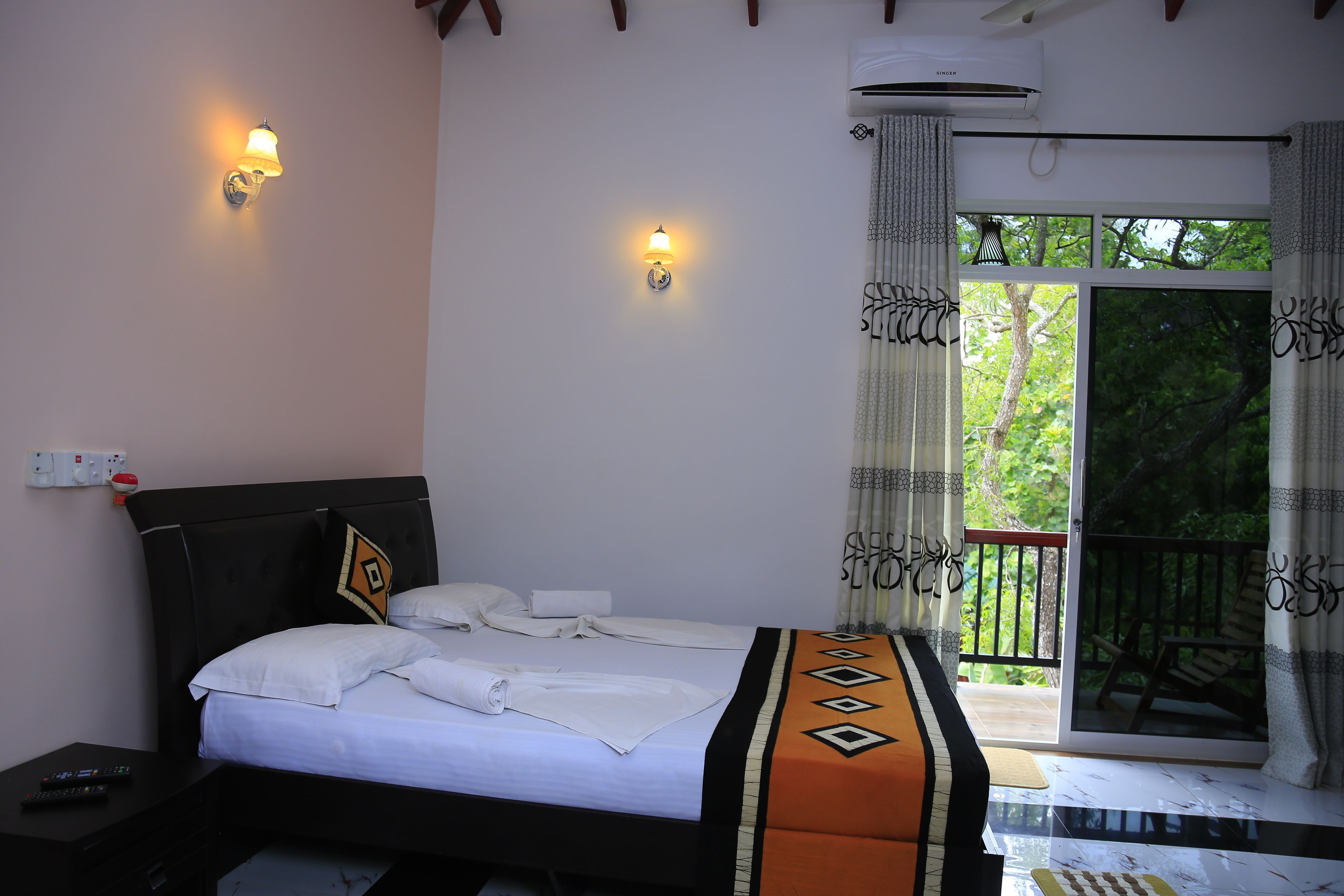 A neatly made bed with white linens and a patterned throw, a nightstand with a remote control, and a balcony with a wooden railing and a view of greenery.