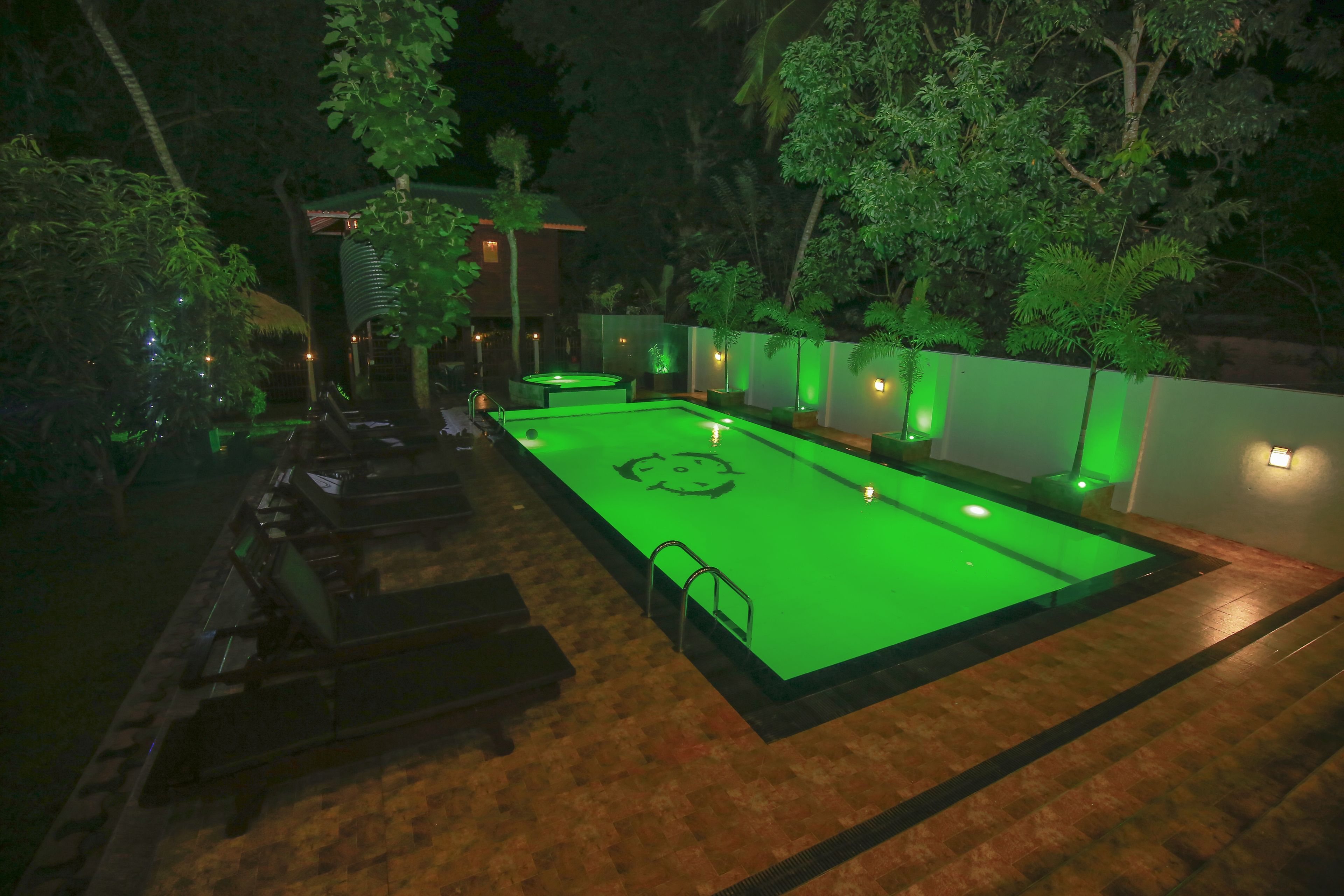 Un'area piscina con un pontile illuminato di verde, circondata da alberi e un edificio con tetto di paglia.