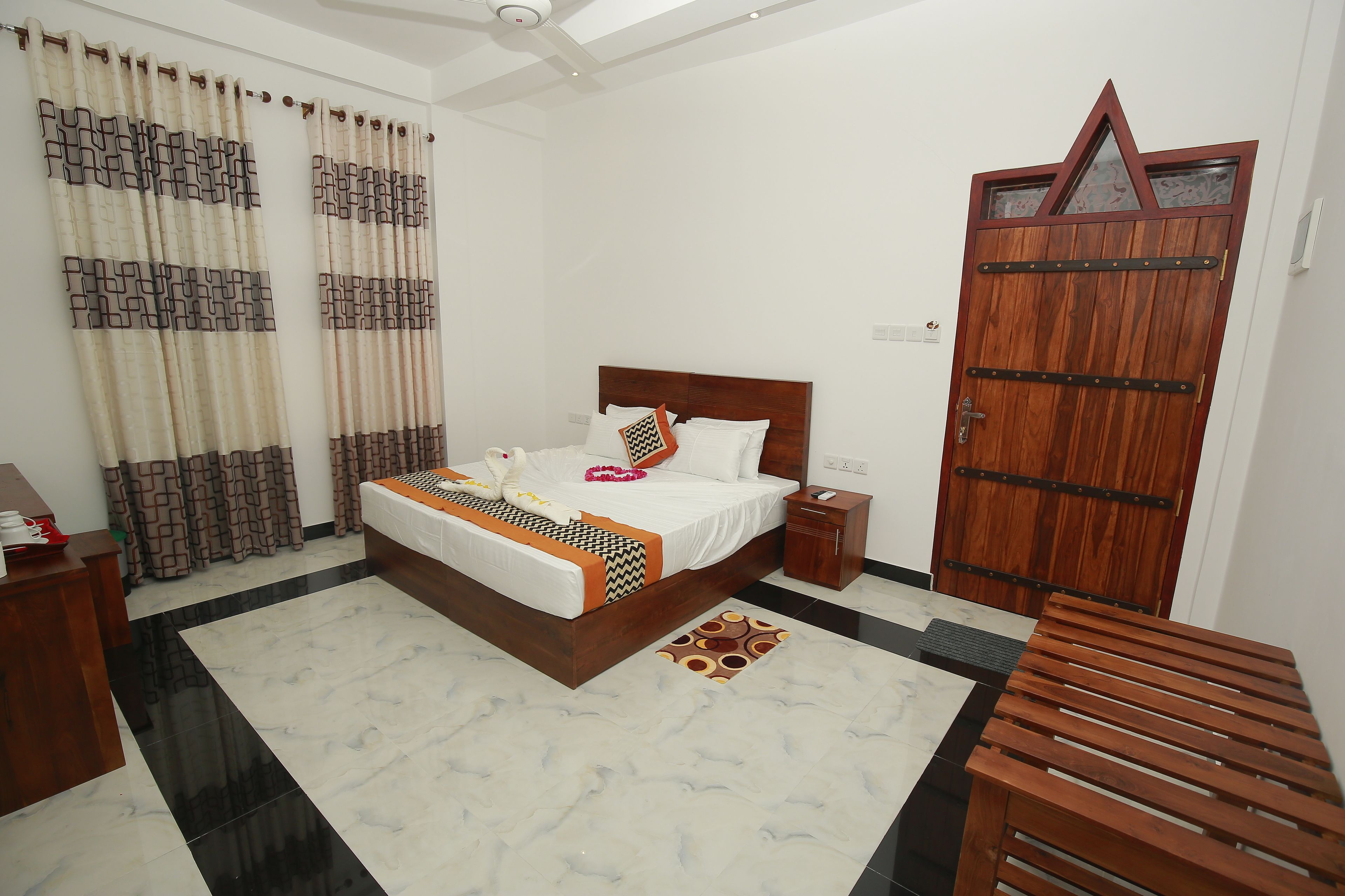 A hotel room with a bed, bedside table, curtains, a wooden door, and a patterned rug.