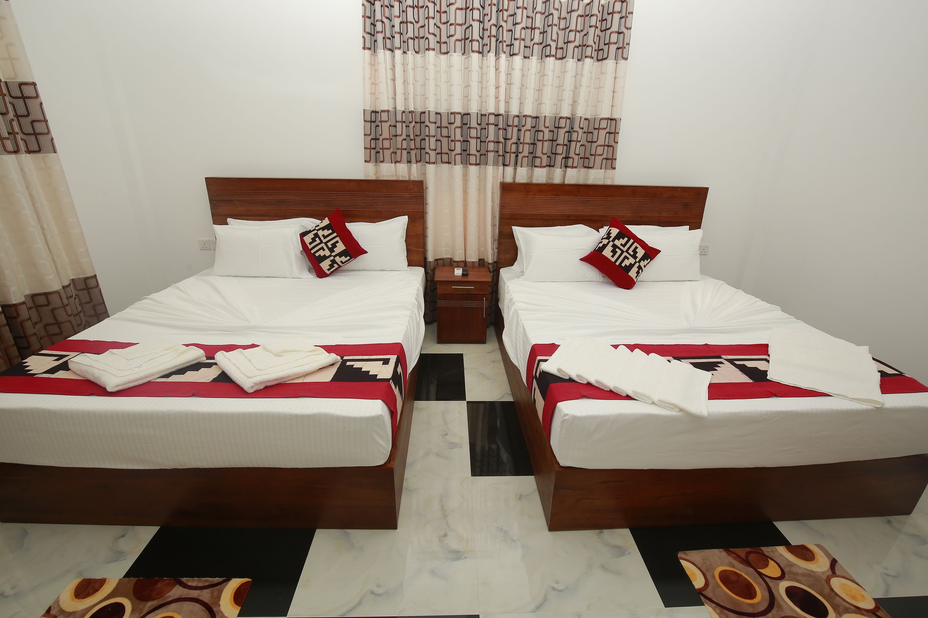 A hotel room with two beds, each with a wooden headboard, white bedding, and red and beige patterned pillows. There is a checkered floor and a patterned rug.