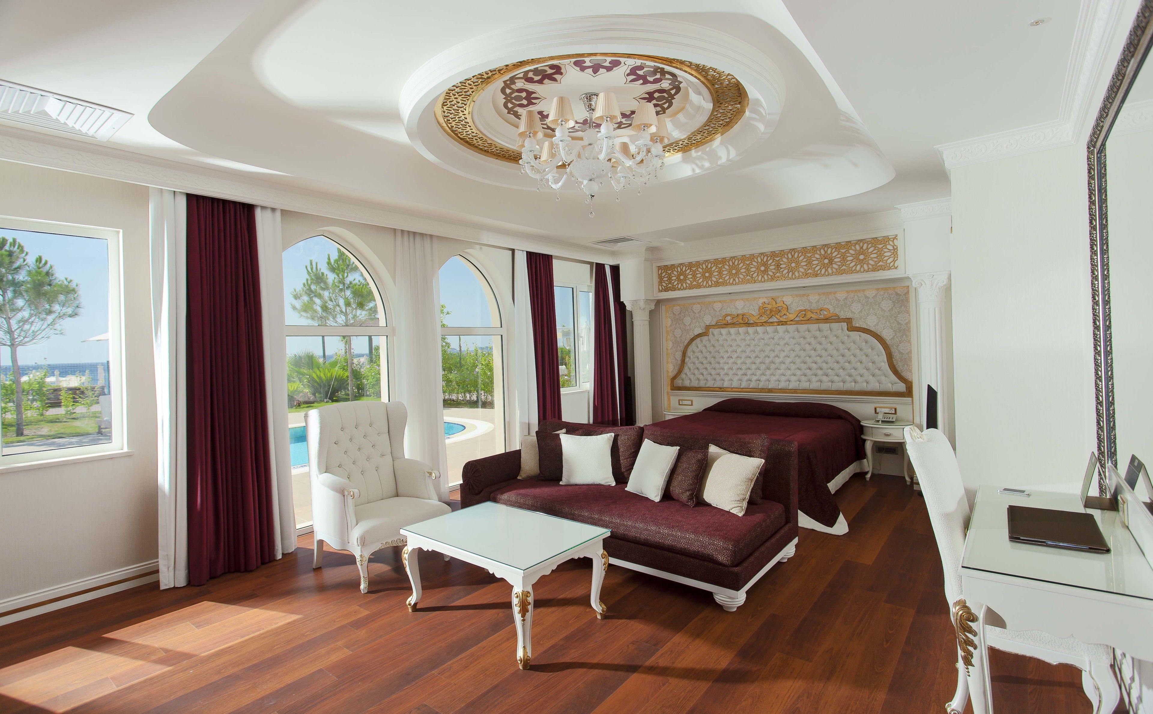 A luxurious hotel room with a large bed, a sitting area with a sofa and armchair, a glass coffee table, and a view of the pool and greenery outside.