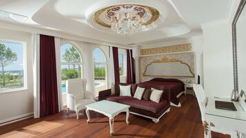 A luxurious hotel room with a large bed, a sitting area with a sofa and armchair, a glass coffee table, and a view of the pool and greenery outside.