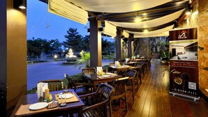Un espacio exterior para comer con mesas y sillas, un letrero y vistas a la calle y los árboles.