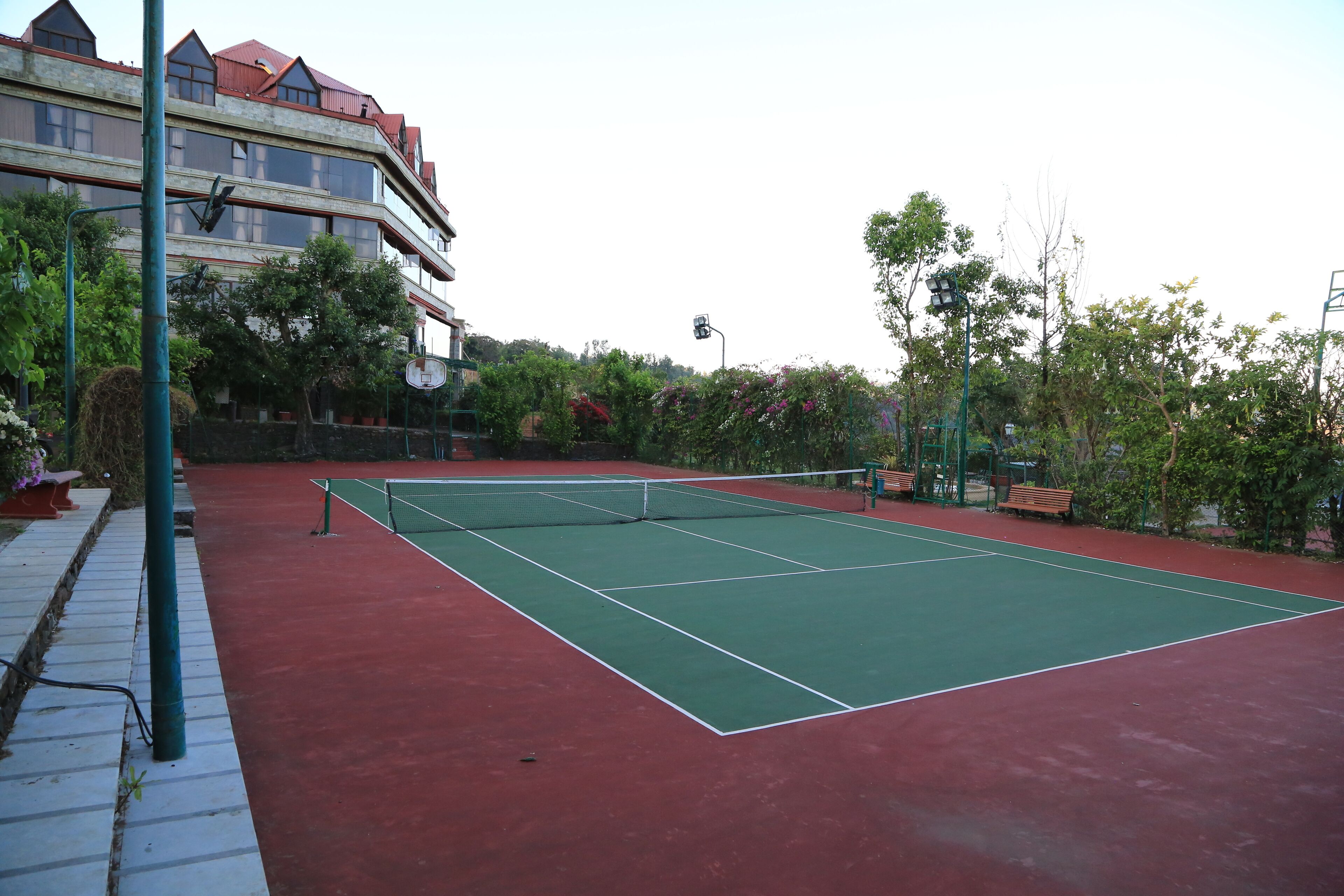 An outdoor tennis court with a red surface, surrounded by greenery and a building with multiple windows in the background.