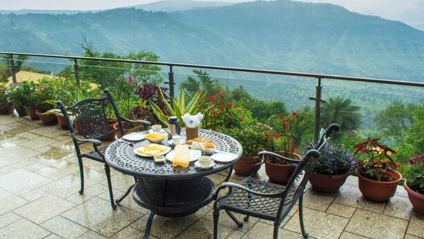 A round metal table with chairs on a balcony, surrounded by potted plants and overlooking a mountainous landscape. - Ravine Hotel (Mahabaleshwar)
