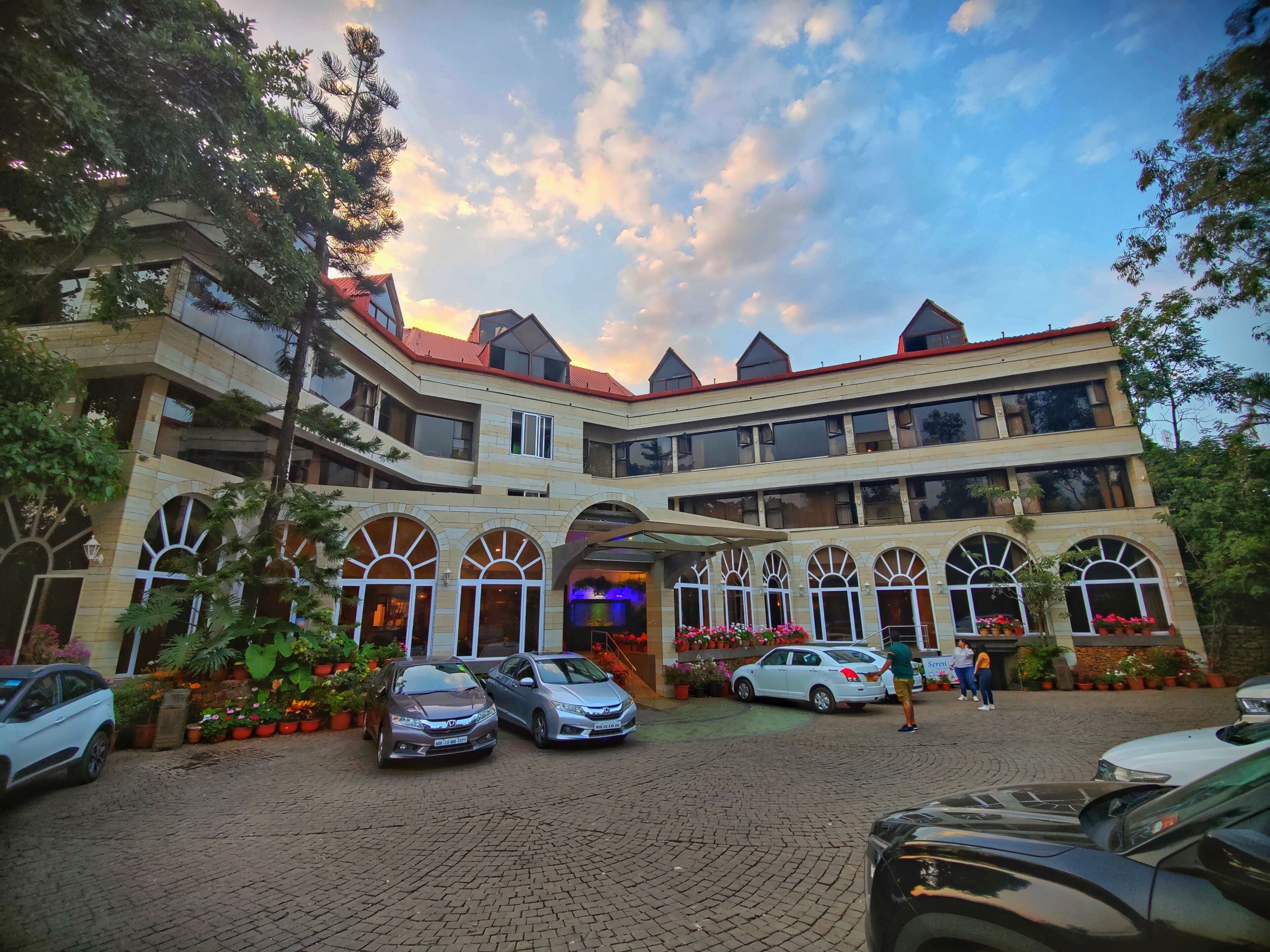 A multi-story hotel with a brick facade, arched windows, and a red roof. There are several cars parked in front, and a person is visible near the entrance.