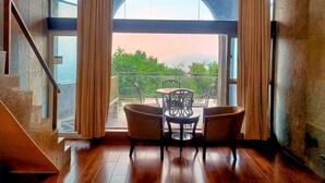 A room with a wooden floor, a table with chairs, and a view through large windows. - Ravine Hotel (Mahabaleshwar)