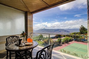 A balcony with a table set for breakfast, a tennis court, and a mountainous landscape view.