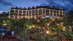 A multi-story hotel with a symmetrical facade, multiple balconies, and a landscaped garden area with a fountain. - Ravine Hotel (Mahabaleshwar)