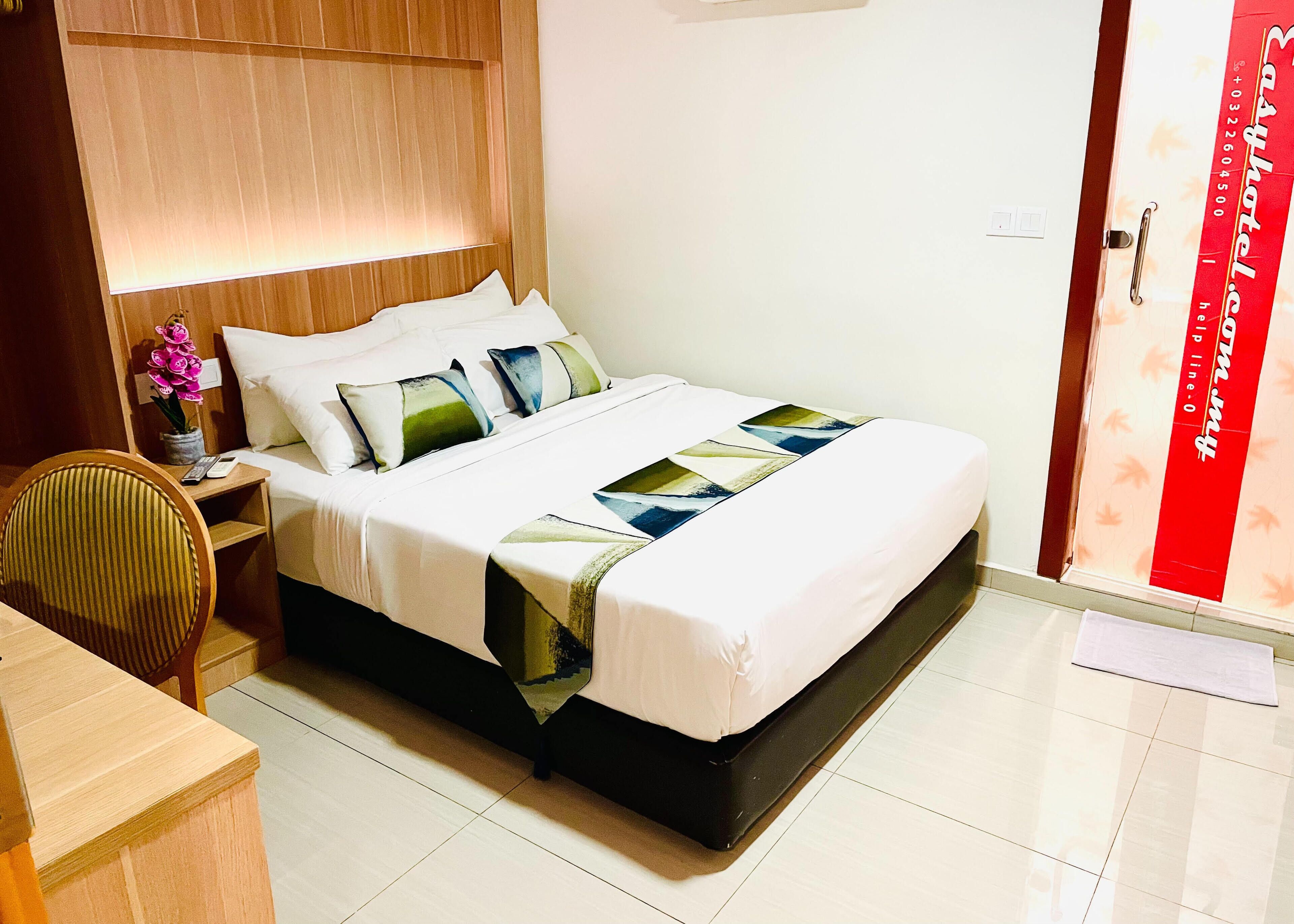 A hotel room with a bed, bedside table, chair, and a red banner on the door.