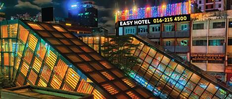 A brightly lit hotel with a modern architectural design, featuring a prominent glass facade and a digital sign displaying the hotel's name and contact number.