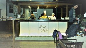 Un restaurante con mostrador, algunos clientes sentados y un cocinero con uniforme.