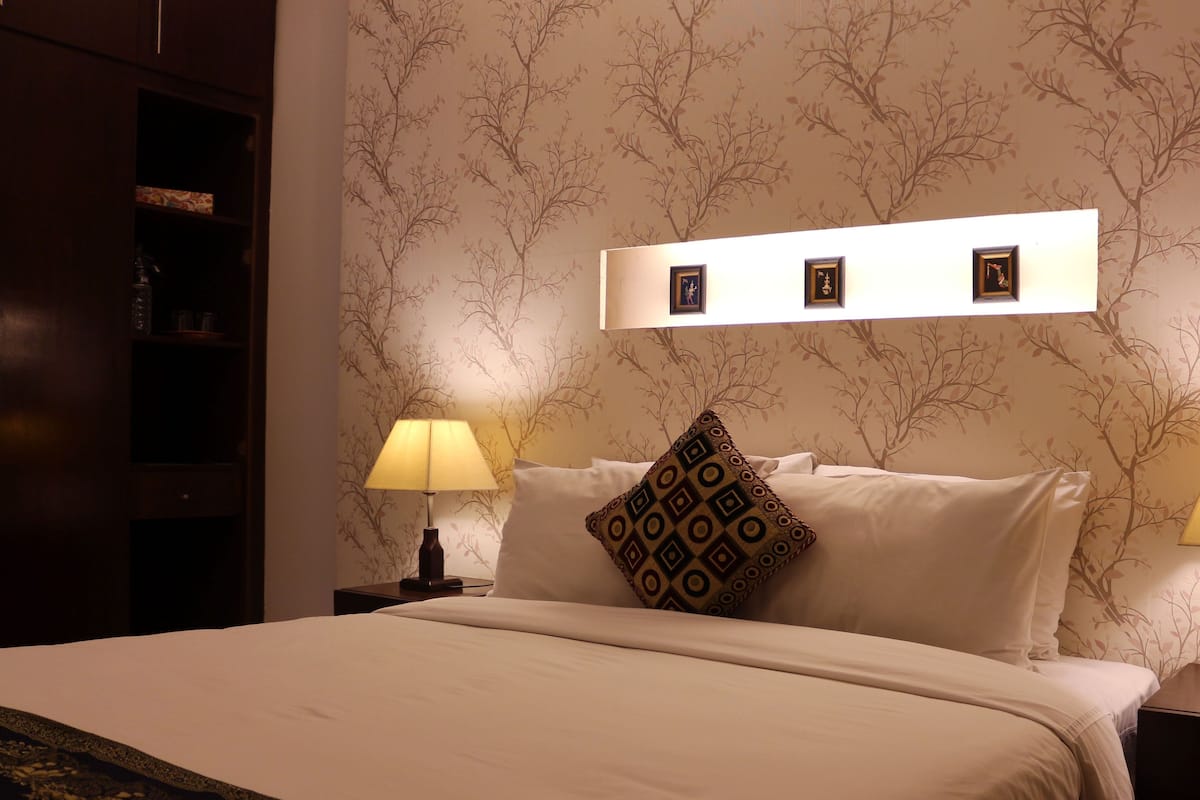 A neatly made bed with a patterned pillow, a bedside lamp, and a wall with framed pictures.
