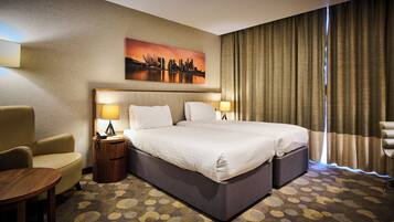Une chambre d’hôtel avec un grand lit, deux tables de chevet avec des lampes, une chaise, une petite table et un tableau représentant un paysage urbain accroché au mur.