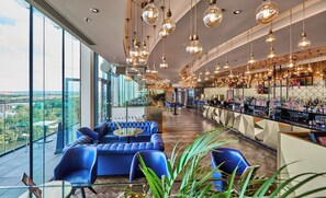Una zona de bar con techo alto y lámparas colgantes, un área de estar con sillas azules y vistas a través de amplios ventanales.