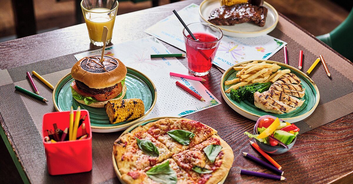 A table with a burger, pizza, grilled chicken, fries, and a salad. There are drinks and pencils on the table.