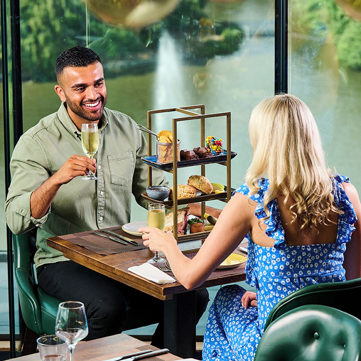 A dining area with a table set for two, a tiered dessert stand, and a view of a body of water through a window.