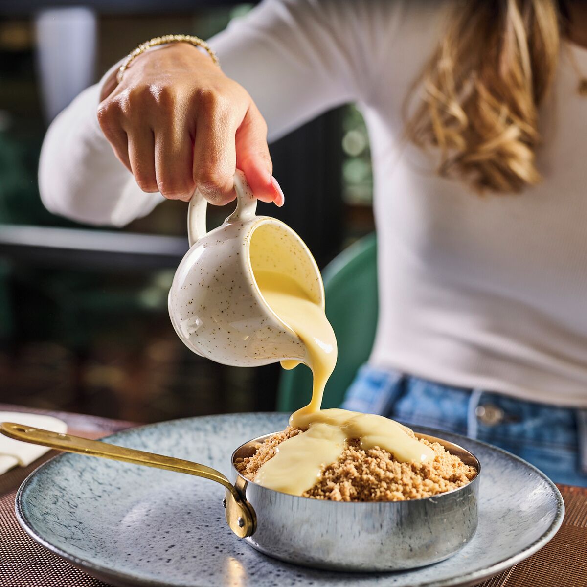 A person pouring a creamy sauce over a dish in a metal bowl on a plate.