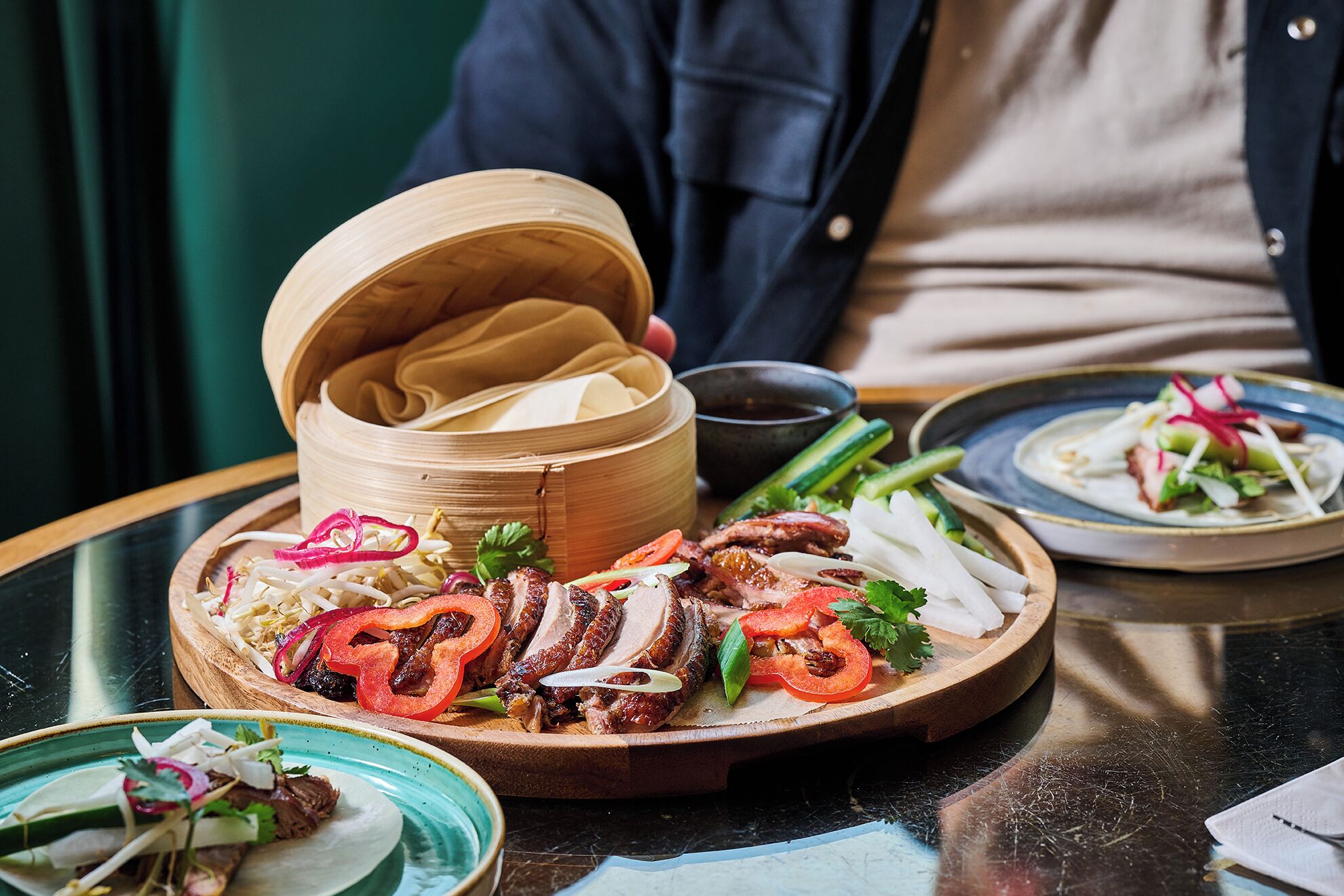A dining table with a variety of dishes, including sliced meat, vegetables, and a bamboo steamer.