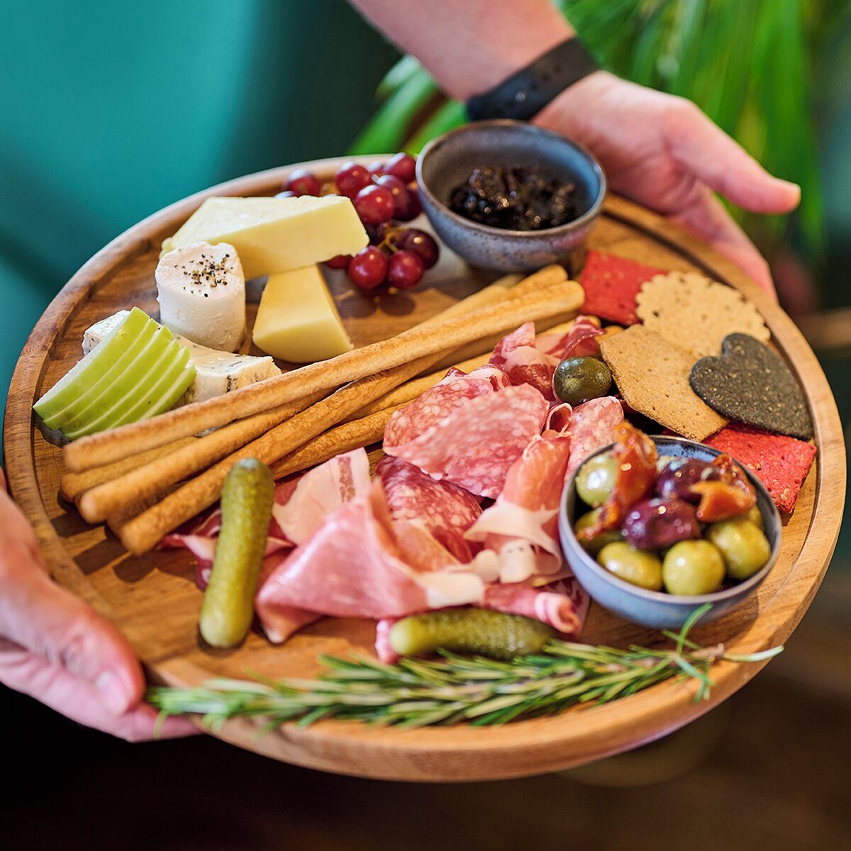 A wooden board with various cheeses, cured meats, olives, and crackers.
