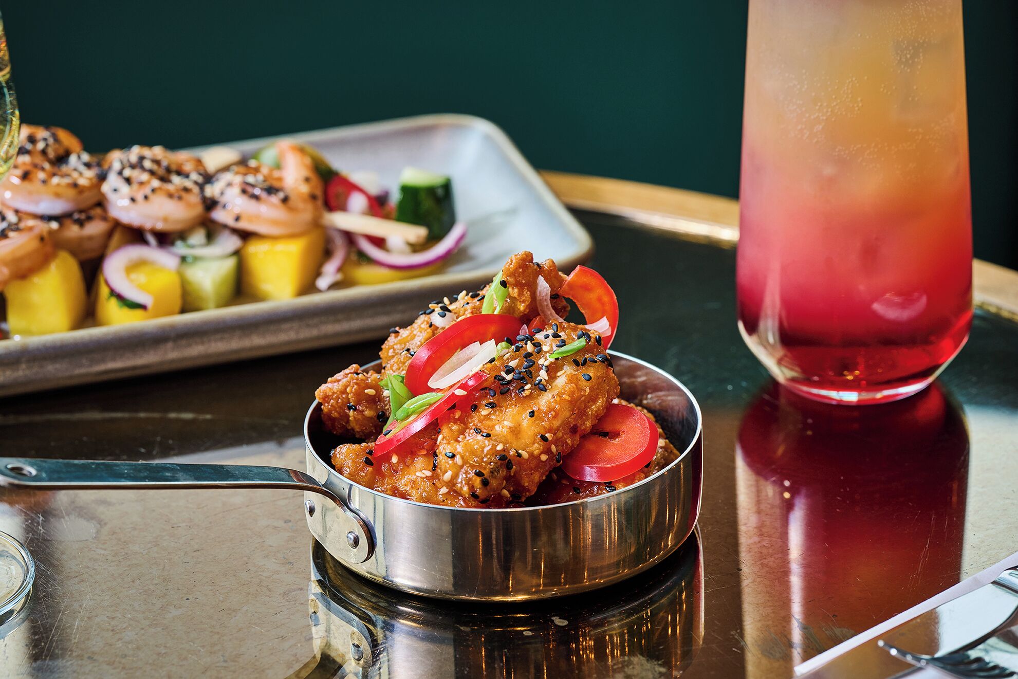 A metal bowl of chicken wings garnished with sesame seeds and sliced vegetables, a tray of assorted pastries, and a glass of iced beverage on a reflective surface.