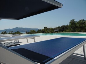 A poolside lounge chair with a blue cushion, a white table, and a view of a clear blue pool with mountains in the background.