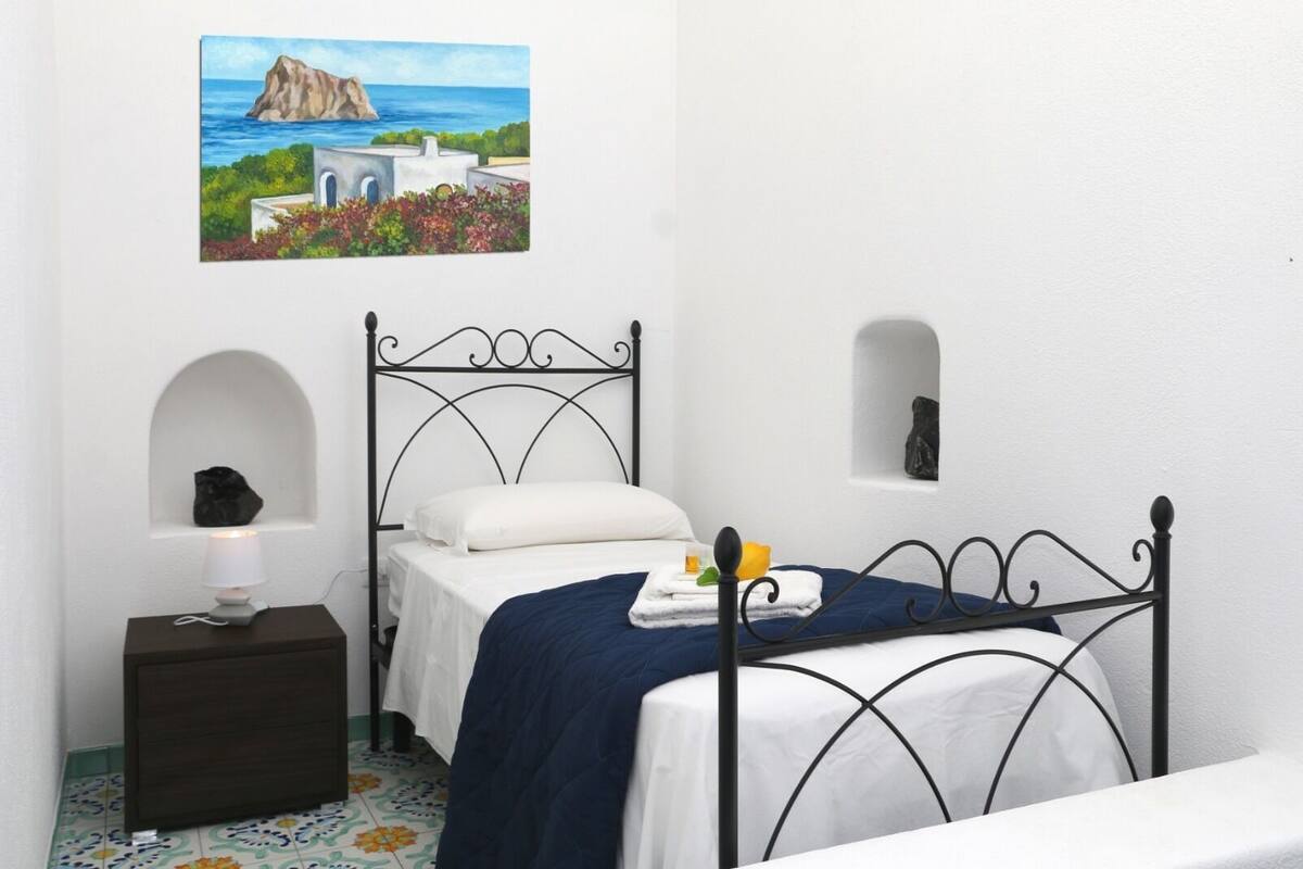 Een slaapkamer met een bed, een nachtkastje, een lamp en een schilderij van een kustlandschap.