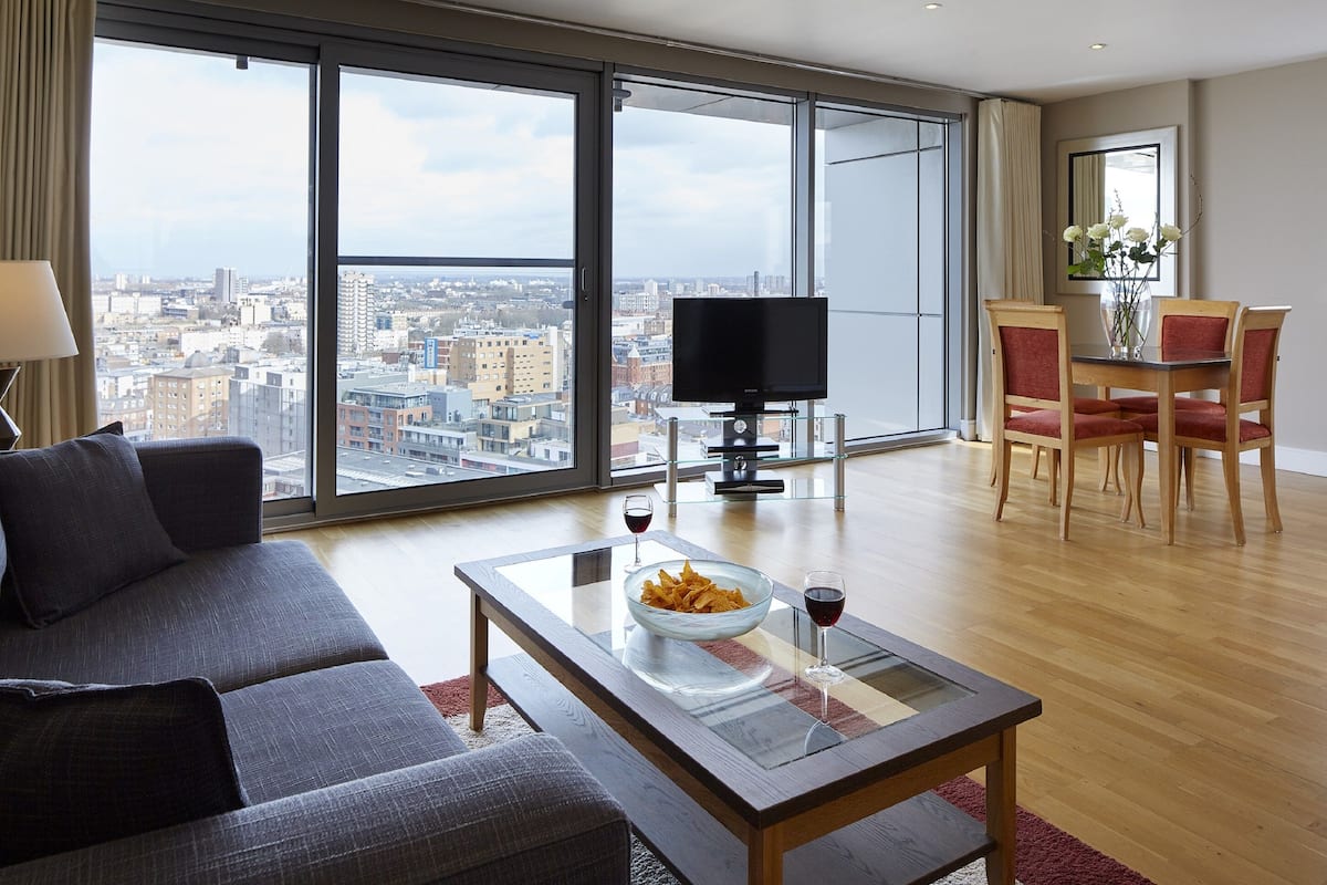 A modern living room with a large window, a flat-screen TV, a dining area with red chairs, and a bowl of snacks on a glass coffee table.