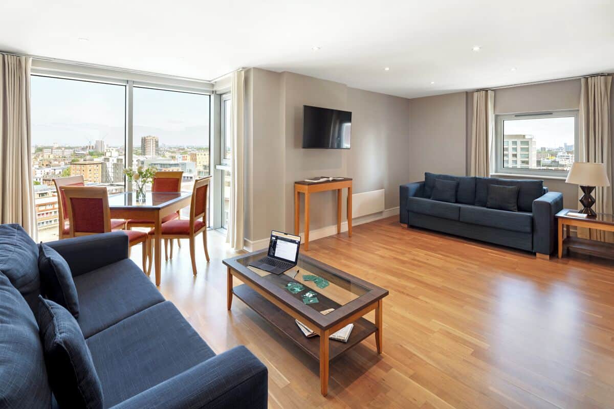 A modern living room with a sofa, a glass coffee table, a TV mounted on the wall, and a view of the city through large windows.