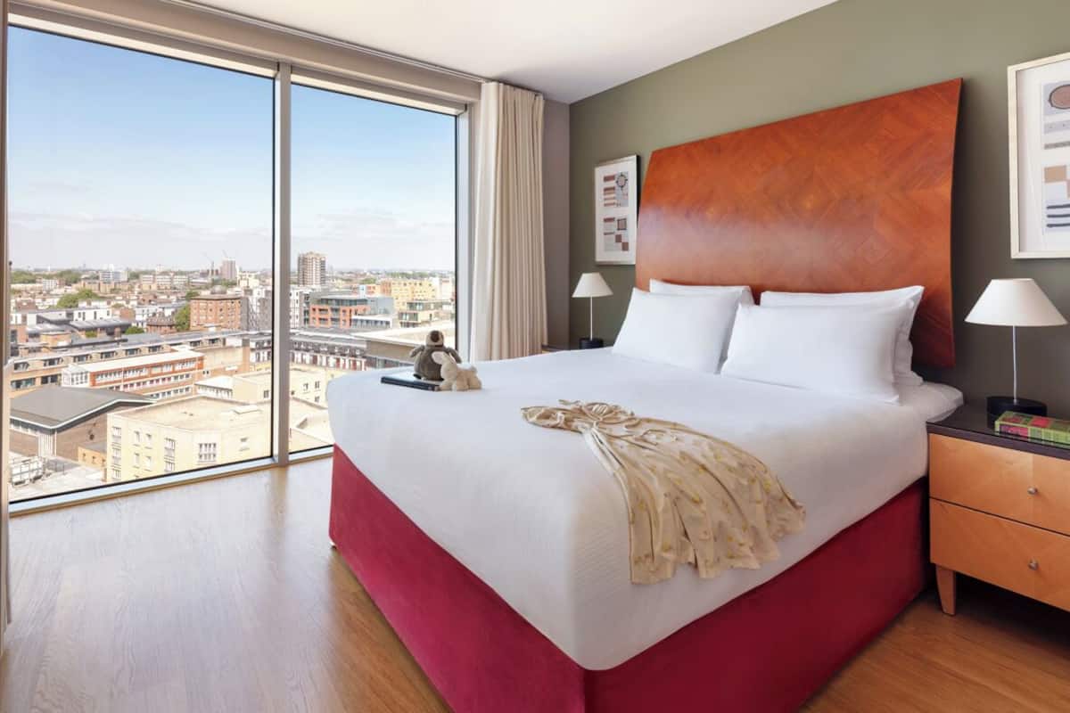 A hotel room with a large bed, a wooden headboard, a nightstand with a lamp, and a view of the city through large windows.