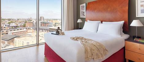 A hotel room with a large bed, a wooden headboard, a nightstand with a lamp, and a view of the city through large windows.