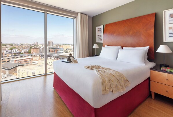 A hotel room with a large bed, a wooden headboard, a nightstand with a lamp, and a view of the city through large windows.