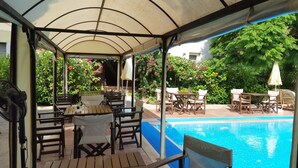 Un área de estar al aire libre con techo, vista a la piscina, muebles de madera y abundante vegetación.