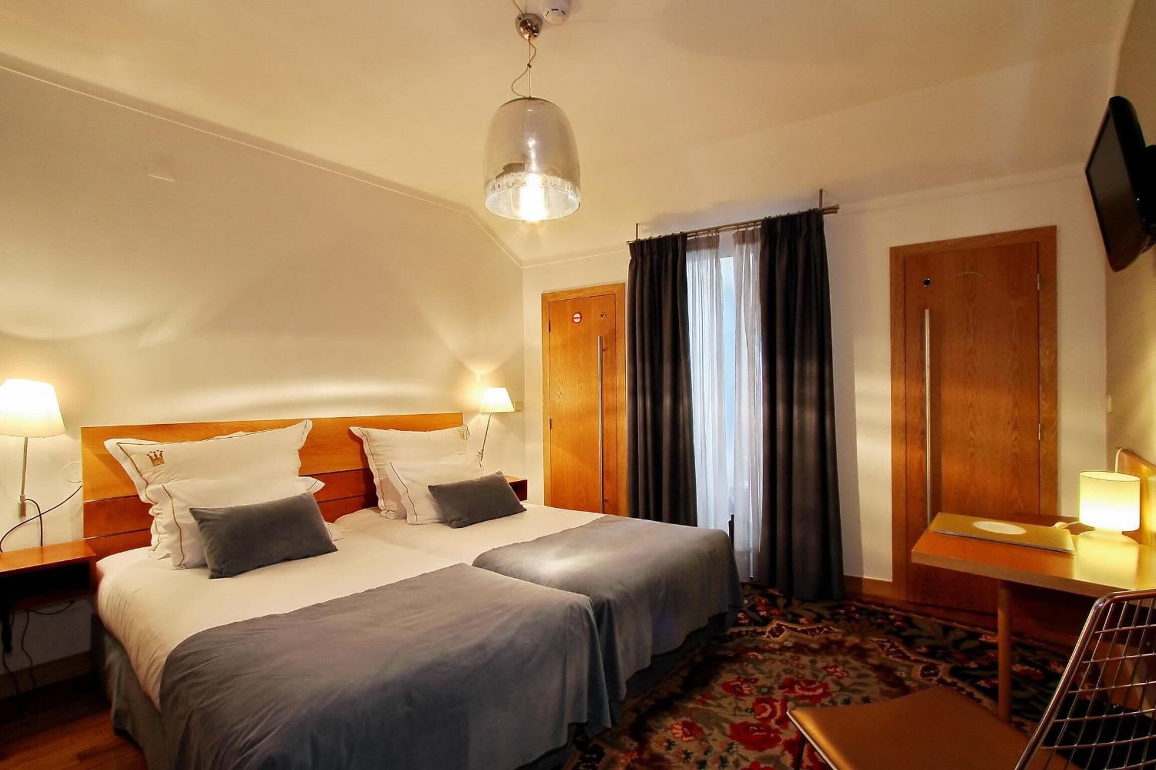 A hotel room with two beds, a wooden headboard, a desk with a lamp, a television, and a patterned carpet.