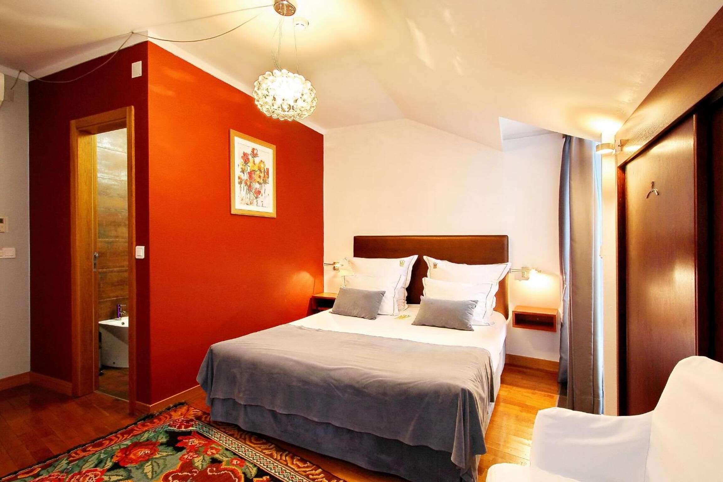 A hotel room with a bed, a nightstand, a bedside table, a chair, a door, and a red wall with a framed picture.