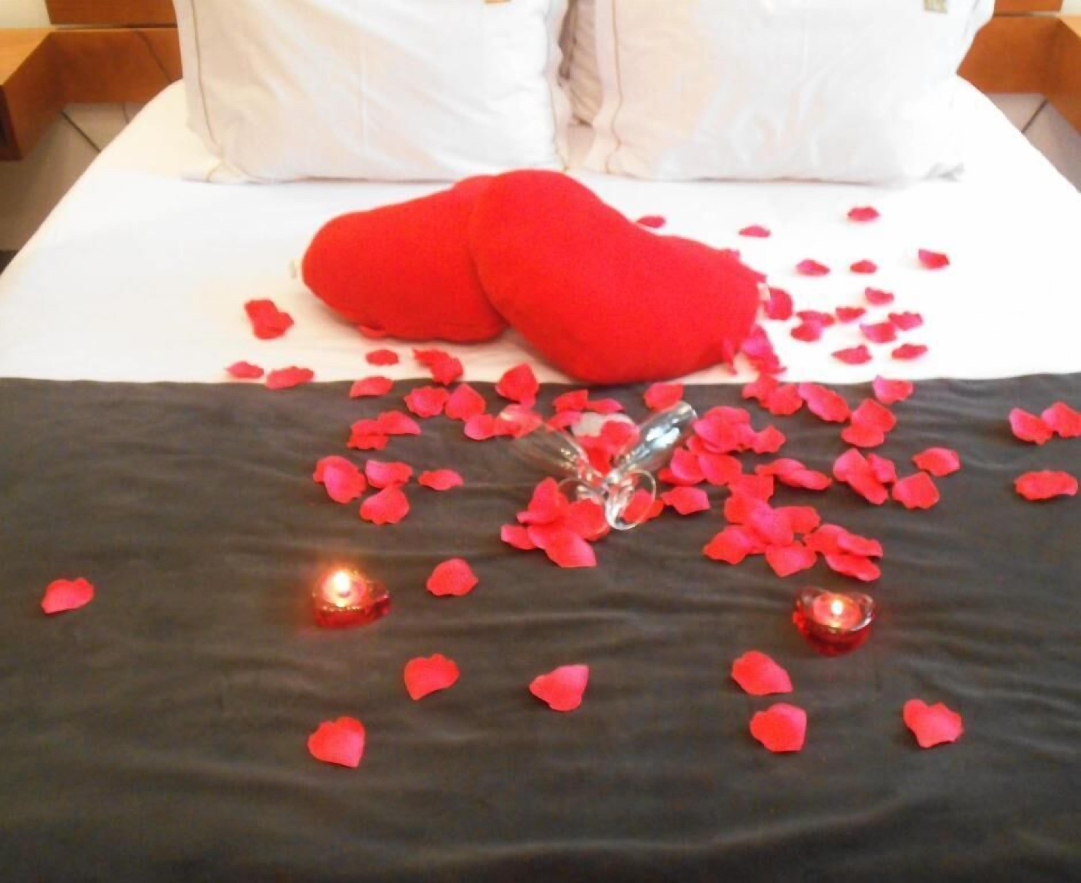 A bed with red rose petals scattered across the bedspread, two red heart-shaped pillows, and two lit candles.