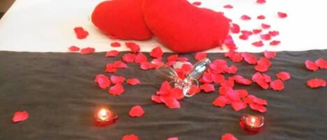 A bed with red rose petals scattered across the bedspread, two red heart-shaped pillows, and two lit candles.
