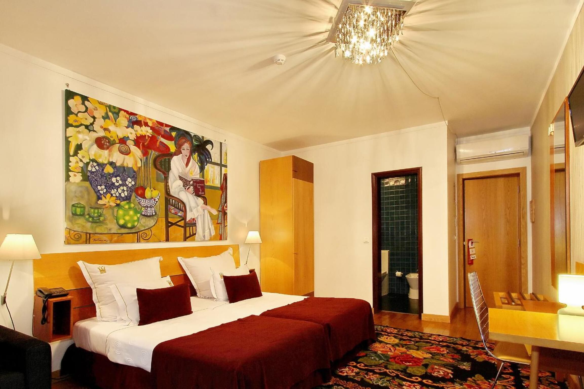 A hotel room with a large bed, two bedside tables, a chandelier, a painting on the wall, and a bathroom visible through an open door.