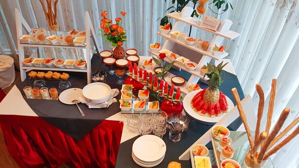 Una mesa de buffet con diversos alimentos y bebidas, dispuesta en varios niveles y acompañada de elementos decorativos como flores y frutas.