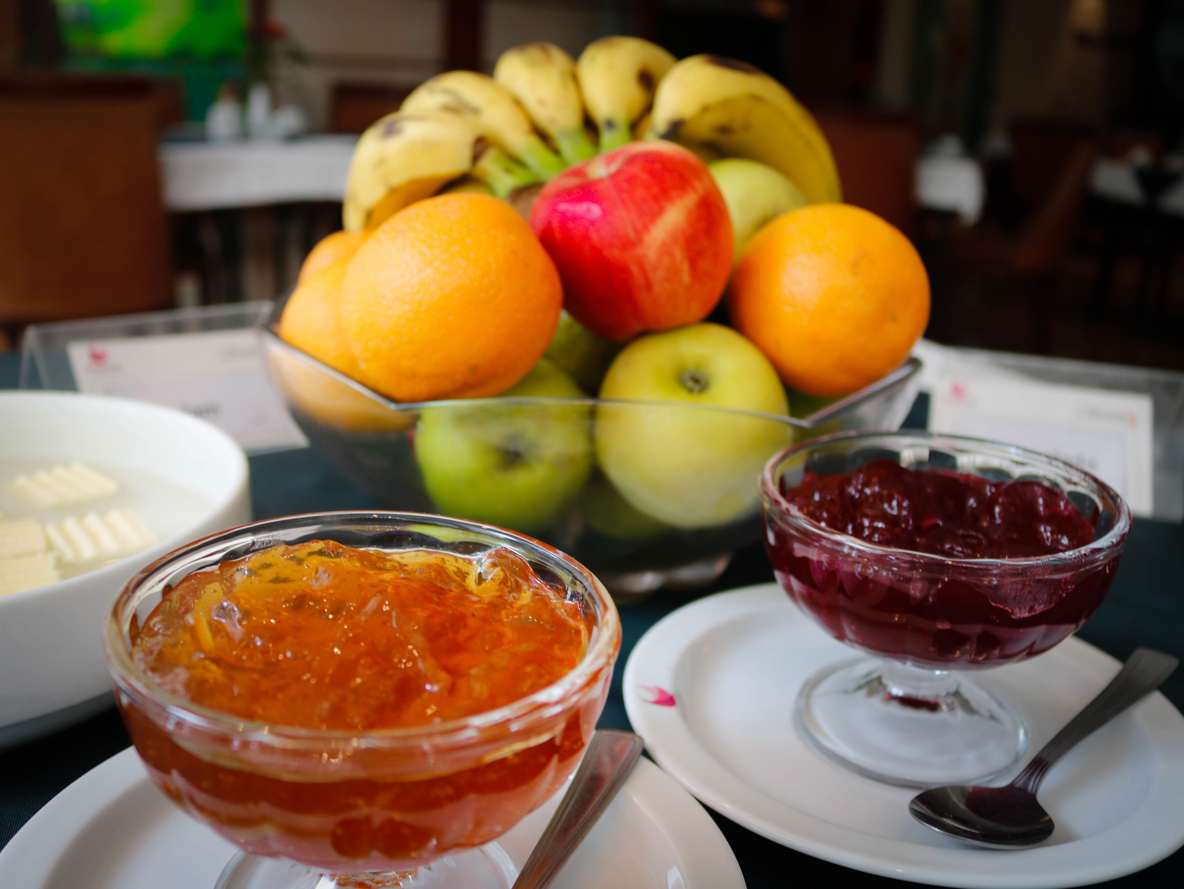 Taula amb un plat de fruita que conté plàtans, pomes i taronges, i dues safates de melmelada de fruita.