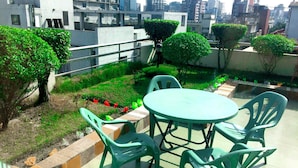 Una terraza en la azotea con una mesa redonda y sillas de plástico verde, rodeada de macetas con plantas y una pequeña zona ajardinada.