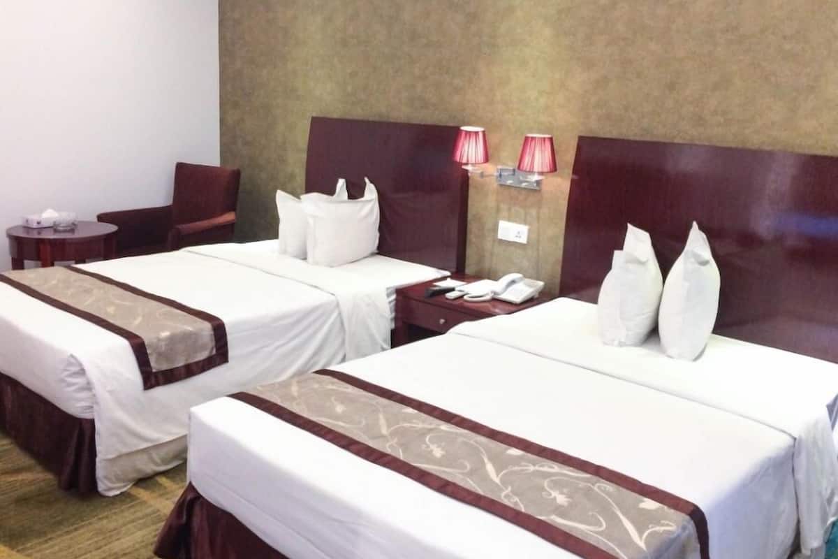 Ein Hotelzimmer mit zwei Betten, jeweils mit weißen Laken und braunen Zierstreifen. Es gibt einen kleinen Tisch mit einer Lampe und einem Sessel, sowie ein an der Wand befestigtes Telefon.