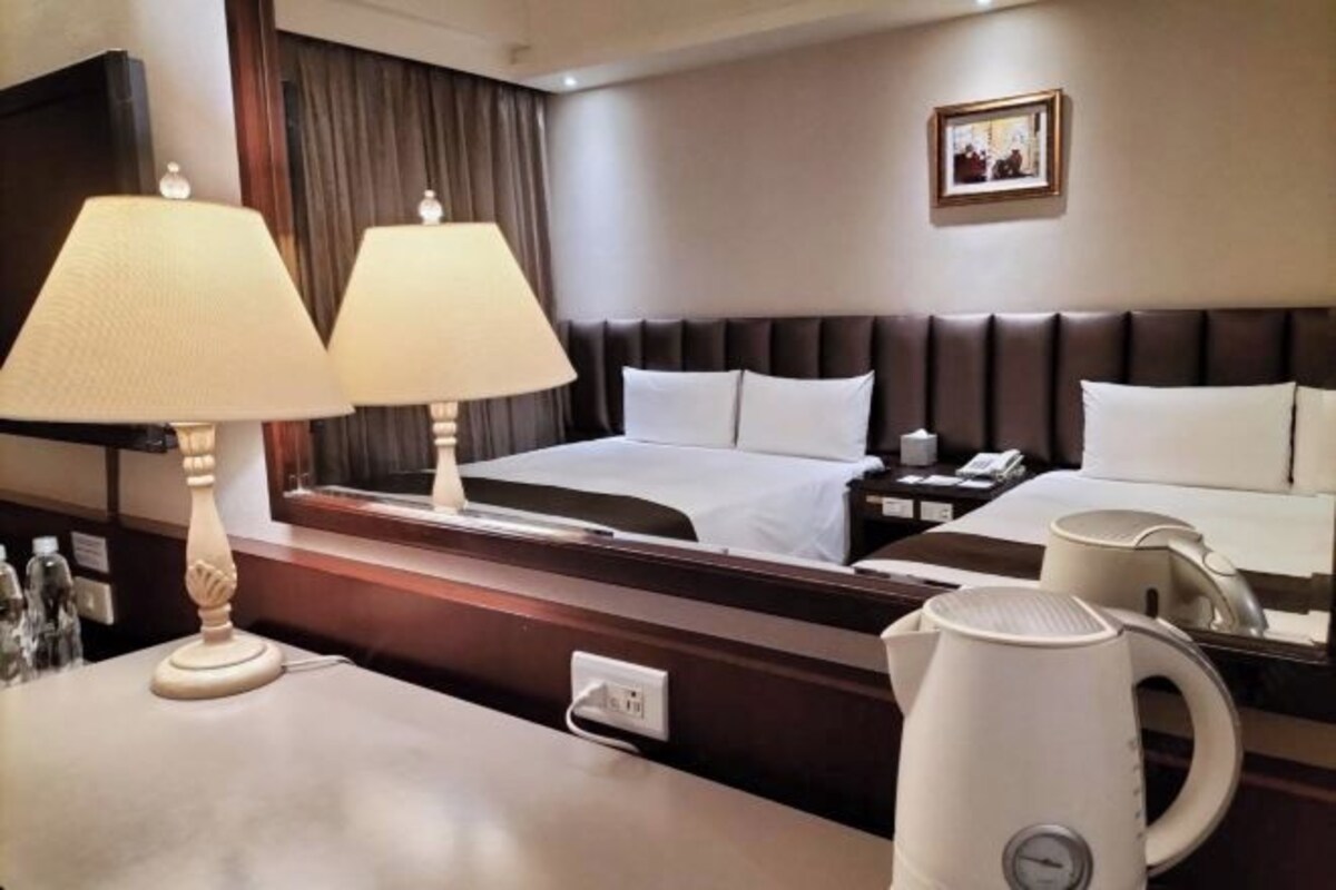 A hotel room with a bed, bedside tables, a lamp, a kettle, and a mirror.