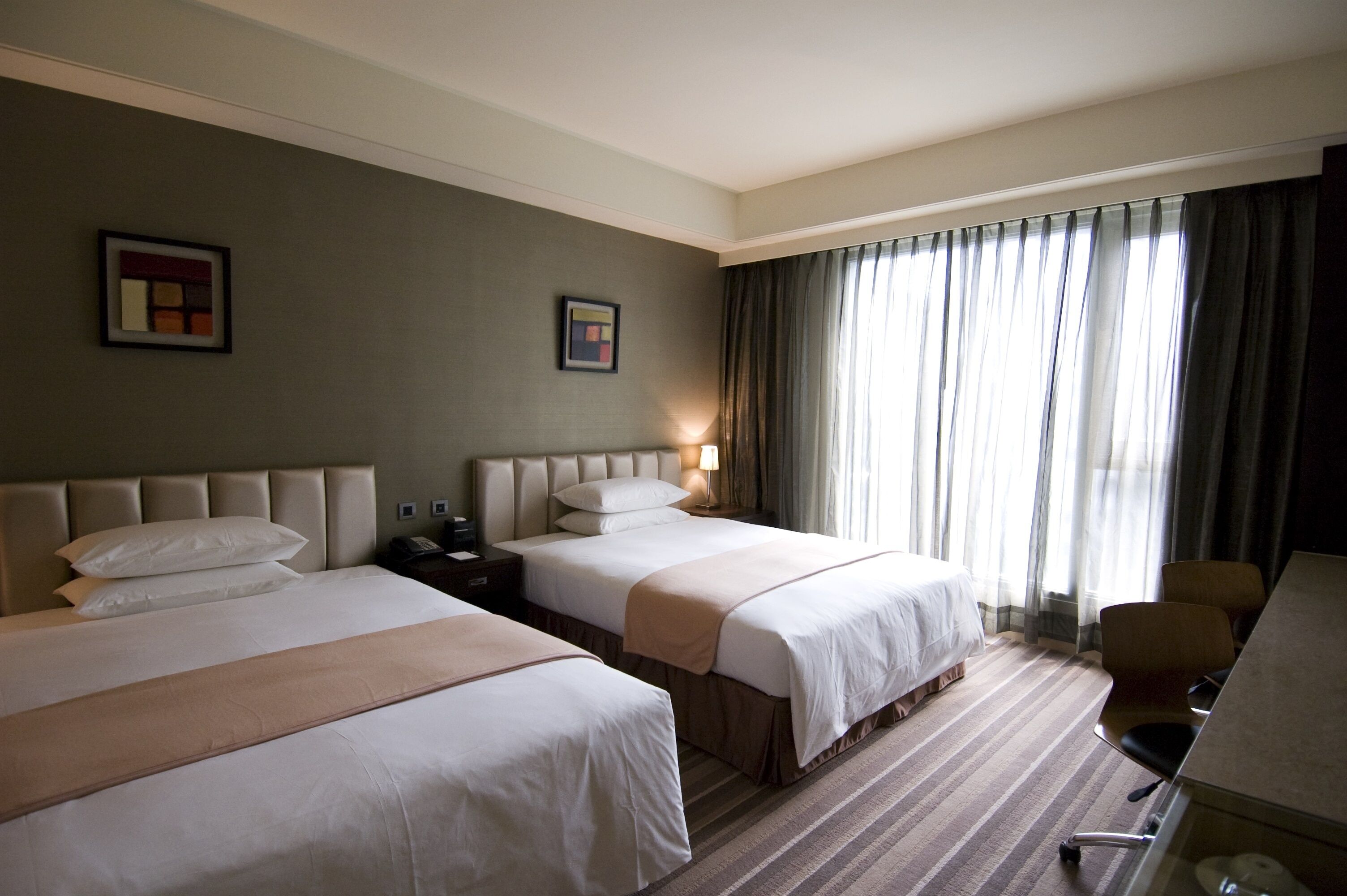 A hotel room with two beds, a desk, and a chair. There are two bedside tables with lamps, a window with curtains, and a framed picture on the wall.