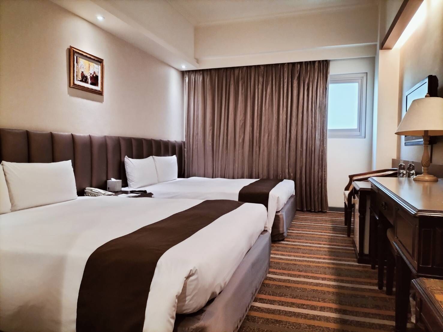 A hotel room with two beds, a desk, a chair, a lamp, and a window with curtains.