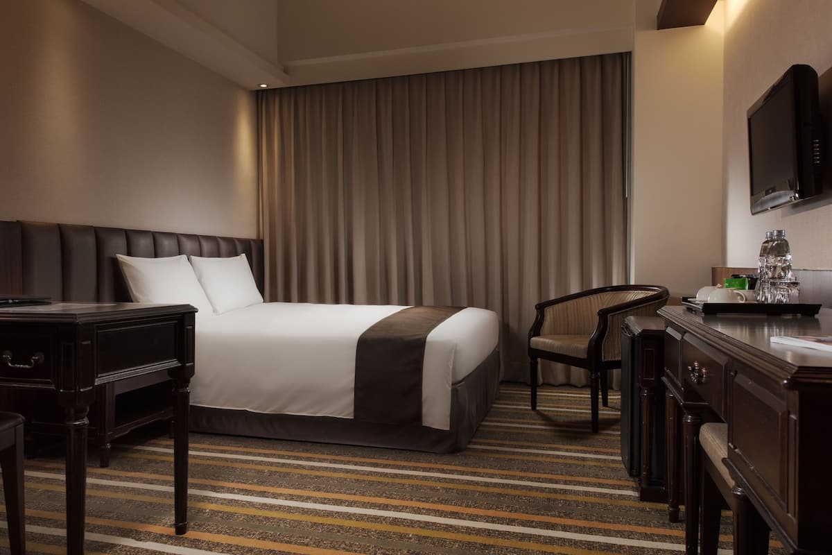 Ett hotellrum med en säng, ett skrivbord, en stol, en lampa, en TV och en minibar.