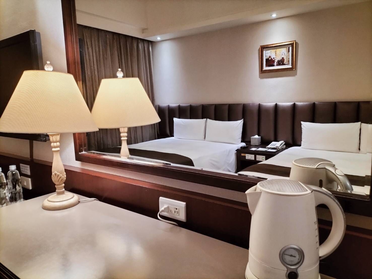 Una habitación de hotel con cama, lámparas de noche, mesita de noche, espejo, un cuadro en la pared y cafetera.