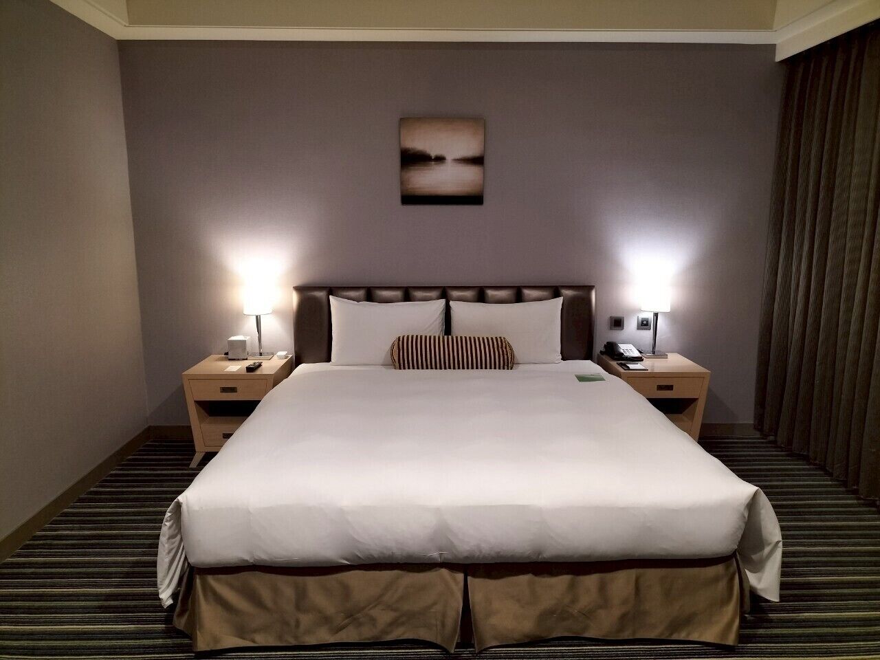 A hotel room with a large bed, two bedside tables with lamps, a nightstand, a wall-mounted picture, and a striped carpet.