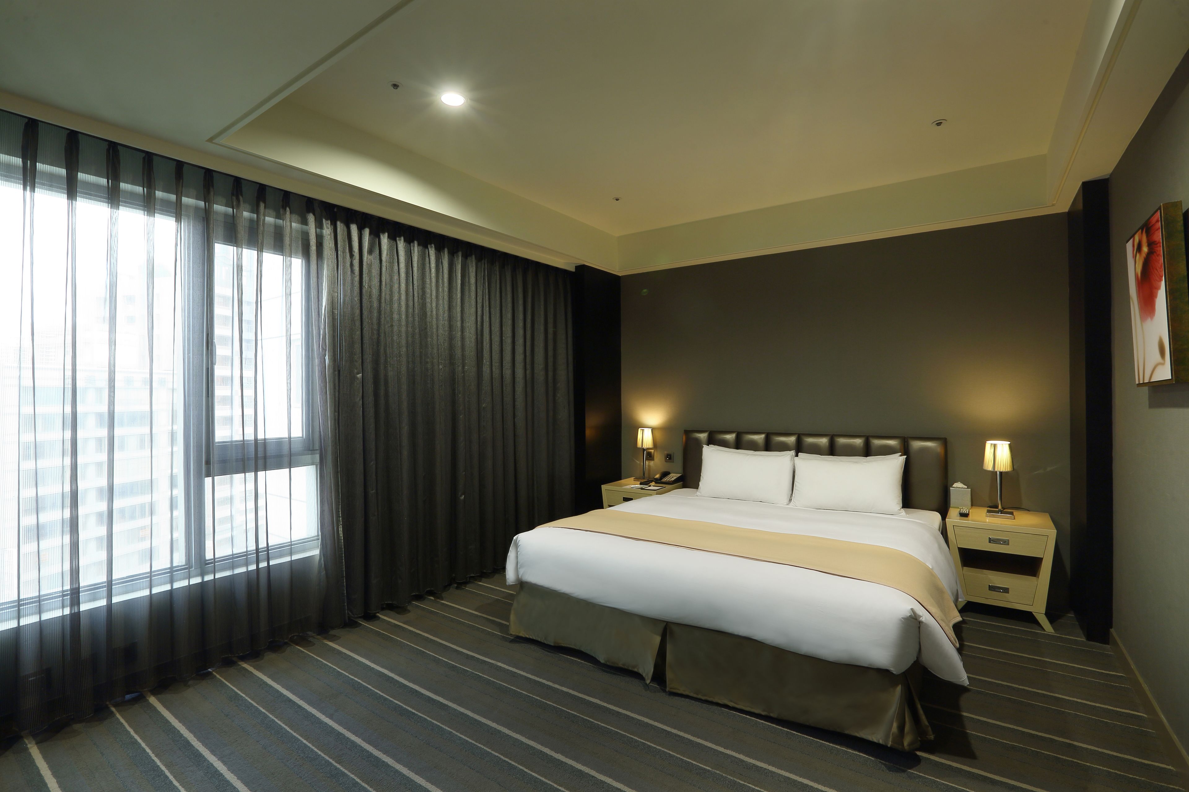 Quarto de hotel com uma cama grande, duas mesas de cabeceira, um criado-mudo, uma janela com cortinas e uma obra de arte na parede.